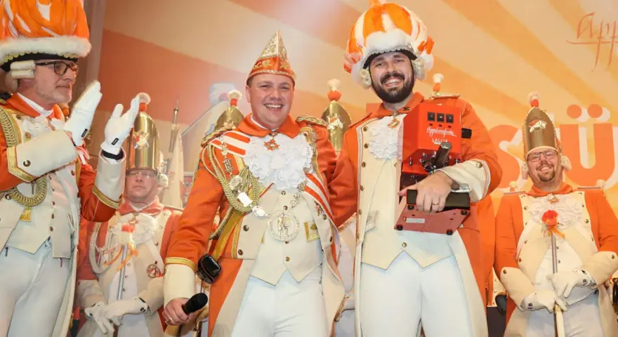 Narren in bunten Kostümen feiern jecke Tage. Ein Mann hält eine Kaffeemaschine, die anderen tragen prunkvolle Uniformen und Federhüte.
