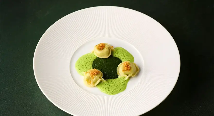Ammolite speisekarte: Drei grüne Ravioli mit goldbrauner Kruste auf einer leuchtend grünen Sauce, angerichtet auf einem weißen Teller.