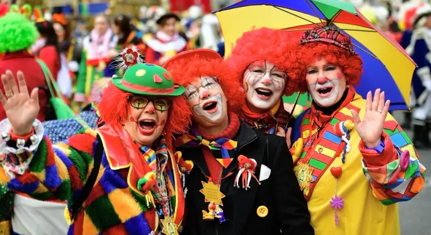 Vier Clowns mit roten Perücken und bunter Kleidung lachen und winken. Ein bunter Regenschirm ist über ihnen. Hier wird kein Karneval gefeiert, sondern ausgelassen gefeiert!