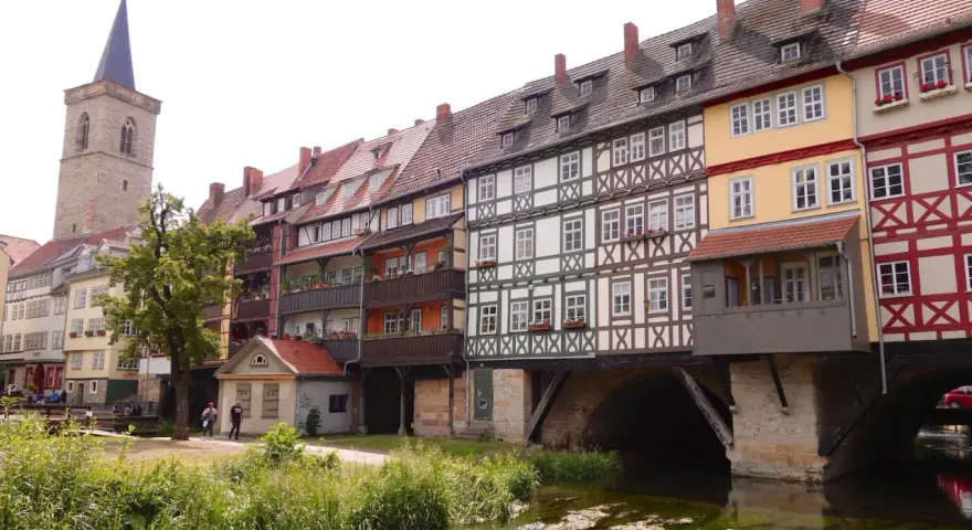 Fachwerkhäuser reihen sich am Wasser entlang, ein Kirchturm ragt empor. Ein idyllischer Anblick, um Erfurt entdecken zu gehen.