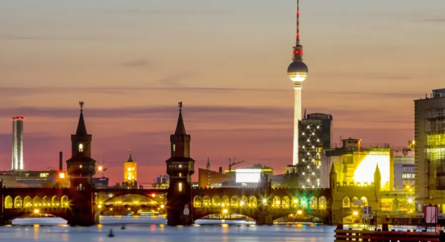 Berlin Kreuzberg bei Sonnenuntergang: Oberbaumbrücke, Fernsehturm und beleuchtete Gebäude spiegeln sich im Wasser.