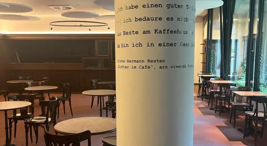 Gemütliches **cafe vegan** mit vielen Tischen und Stühlen. Eine Säule ziert ein Zitat von Hermann Kesten über das Kaffeehaus.