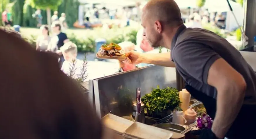 Mann reicht ein Gericht auf einem Holzbrett bei einem Street Food Festival. Die Preise sind hier fair für die leckere Auswahl.