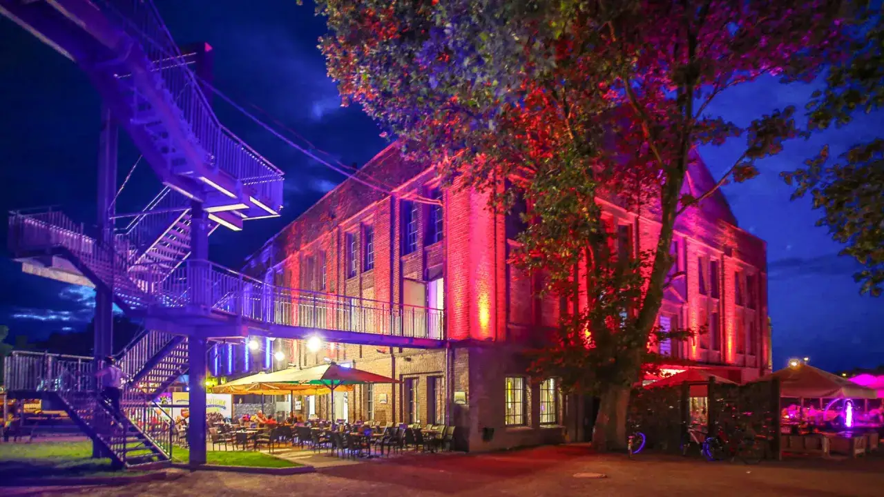 Abendstimmung im Extraschicht Dorsten: Eine beleuchtete Treppe f&uuml;hrt zu einem Geb&auml;ude, das in rotes Licht getaucht ist. Drau&szlig;en laden Tische und St&uuml;hle zum Verweilen ein.
