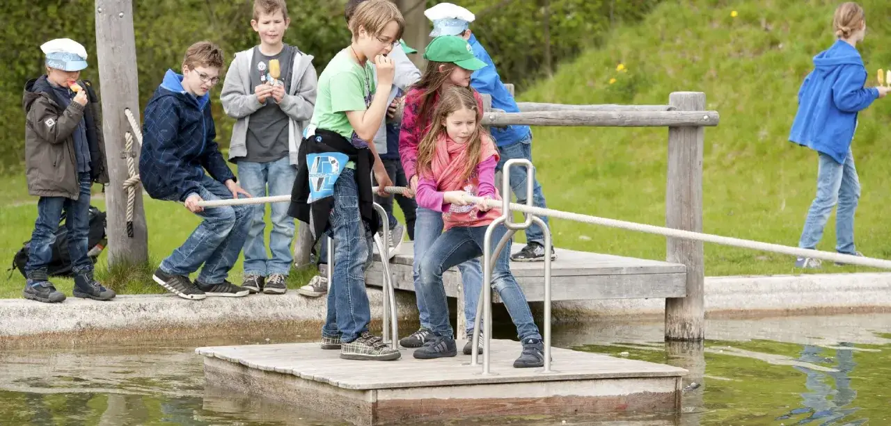 Kinder ziehen an einem Seil auf einem h&ouml;lzernen Steg, der an einen Wasserspielplatz mit Schiffshebewerk erinnert.