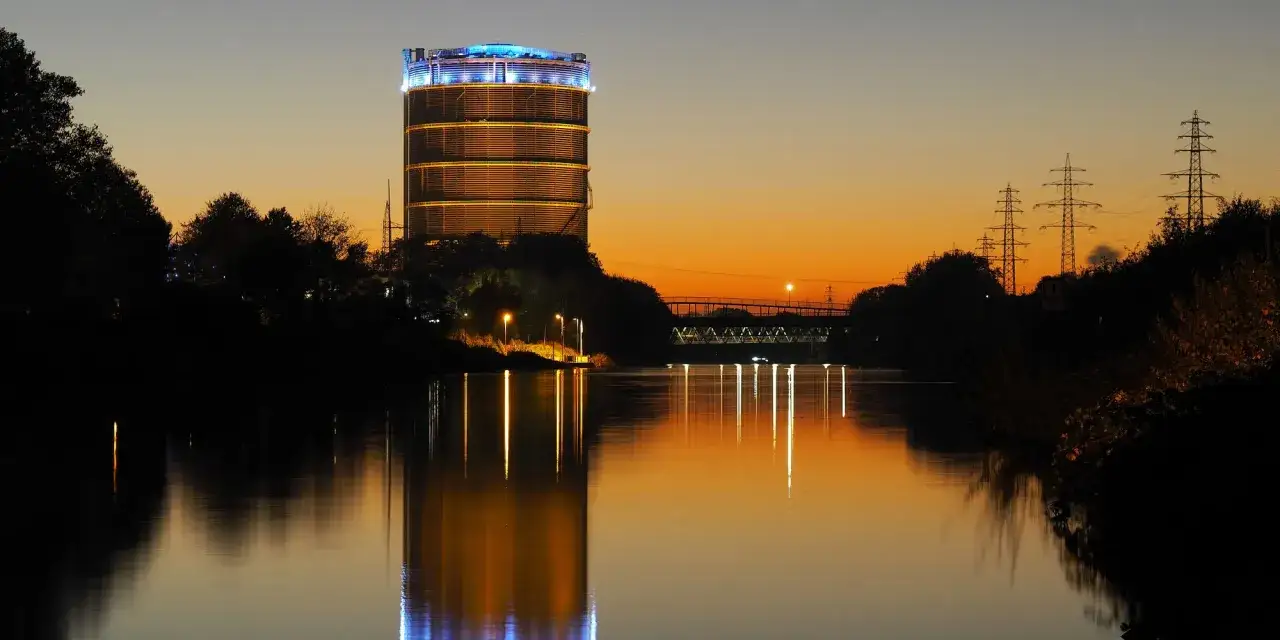 Der beleuchtete Gasometer Oberhausen spiegelt sich im ruhigen Wasser bei Sonnenuntergang.