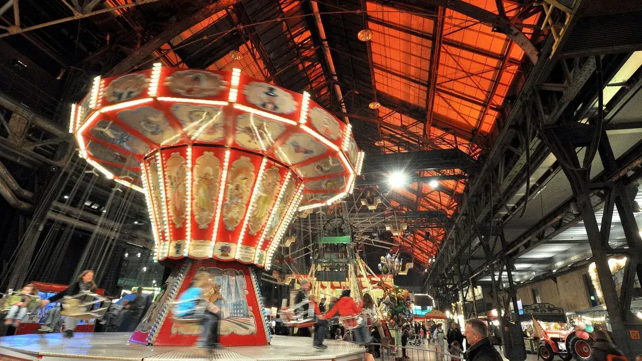 Ein historischer Jahrmarkt in der Jahrhunderthalle mit einem beleuchteten Kettenkarussell, das sich dreht. Menschen genie&szlig;en die Atmosph&auml;re.