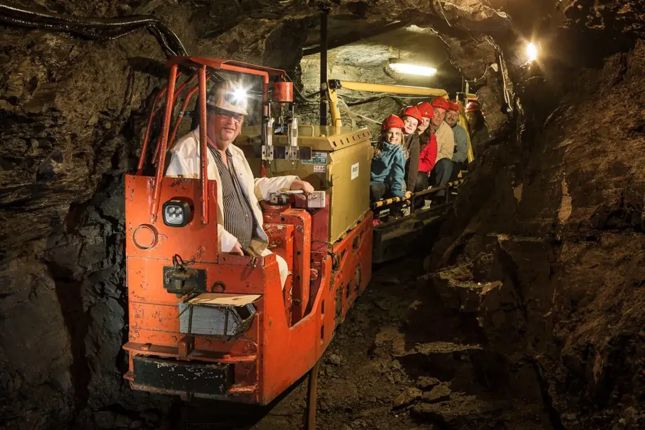 Eine Gruppe von Kindern und Erwachsenen fährt mit einer Lore durch einen dunklen Stollen, um ein Bergwerk zu besichtigen.