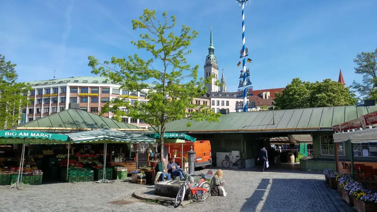 Ein sonniger Tag am Viktualienmarkt in München. Stände mit frischem Obst und Gemüse laden zum Bummeln ein, während im Hintergrund Kirchtürme und ein Maibaum für das typische München-Flair sorgen. Perfekt für munich sightseeing!
