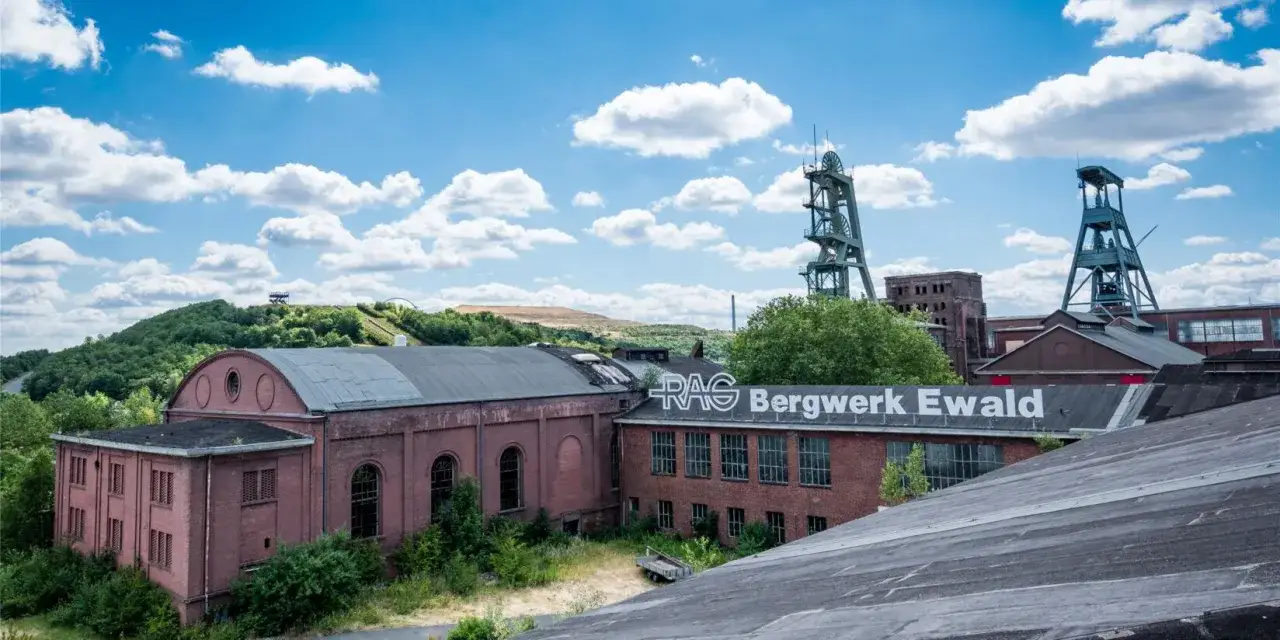 Industriearchitektur des Steelpark Ewald mit F&ouml;rdert&uuml;rmen und Backsteingeb&auml;uden unter blauem Himmel mit Wolken.