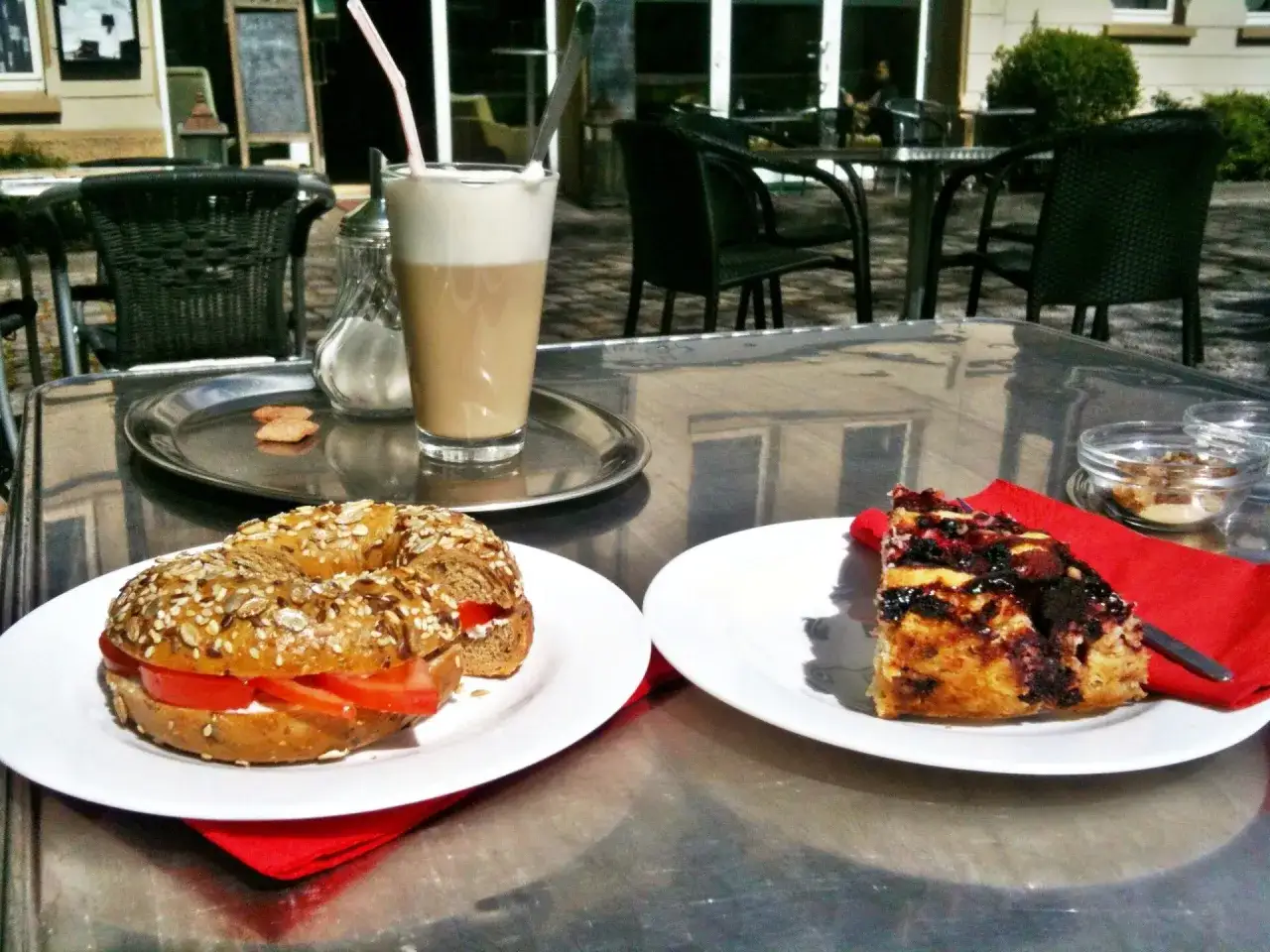 Ein veganes Cafe-Erlebnis: Bagel mit Tomate und Frischkäse, ein Stück Kuchen und ein Latte Macchiato auf einem Tisch im Freien.