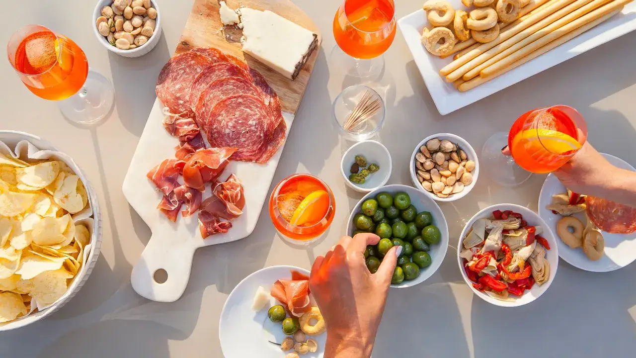 Ein Tisch voller Köstlichkeiten: Aperol Spritz, Salami, Schinken, Käse, Chips, Oliven und Grissini laden zum Genießen ein. Perfekt für eine spritz bar.