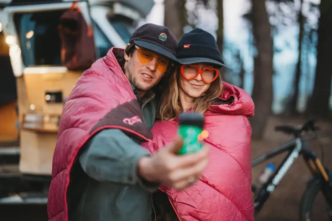 Ein Paar im Freizeithaus, eingewickelt in rosa Decken, macht ein Selfie. Sie tragen lustige Brillen und M&uuml;tzen. Perfekter Freizeitpark-Outfit!
