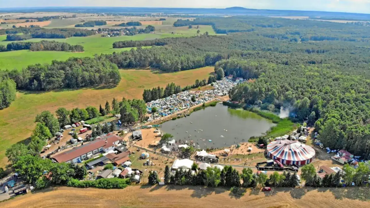 Luftaufnahme eines gro&szlig;en Festivals am See mit Zelten, B&uuml;hnen und einem Zirkuszelt. Das Nachtigall Festival ist in vollem Gange.