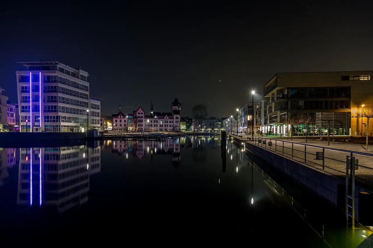 Moderne und historische Gebäude spiegeln sich im ruhigen Wasser. Dortmund bei Nacht, ein malerischer Anblick mit Lichtern, die die Dunkelheit durchbrechen.