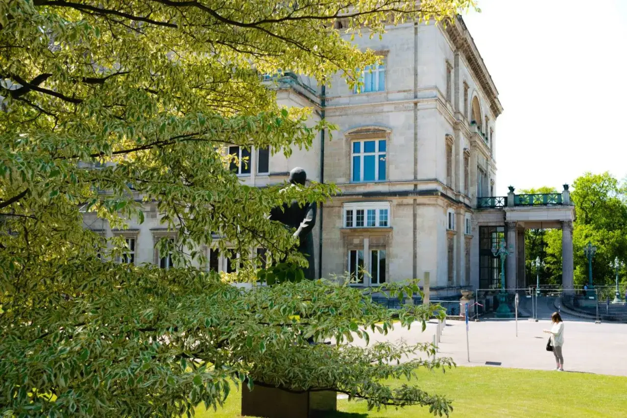 Prächtige Krupp Villa mit Statue im Vordergrund, umgeben von üppigem Grün und einem Baum mit bunten Blättern.