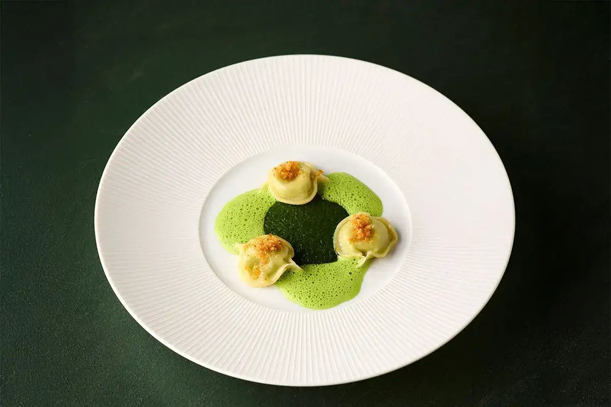 Ammolite Speisekarte: Drei grüne Ravioli mit einer cremigen Kräutersauce und knusprigen Krümeln auf einem weißen Teller.