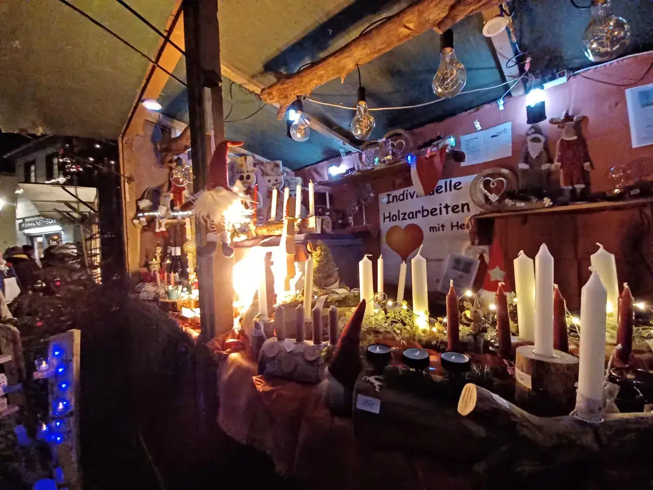 Kerzen, Gnome und Holzdekorationen auf einem Stand beim Monschau Weihnachtsmarkt. Lichterketten und Glühbirnen sorgen für stimmungsvolles Licht.
