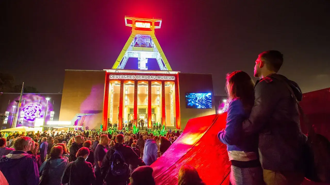 Ein Paar genießt die Extraschicht Bochum vor dem beleuchteten Deutschen Bergbau-Museum. Eine Menschenmenge feiert unter dem Förderturm.