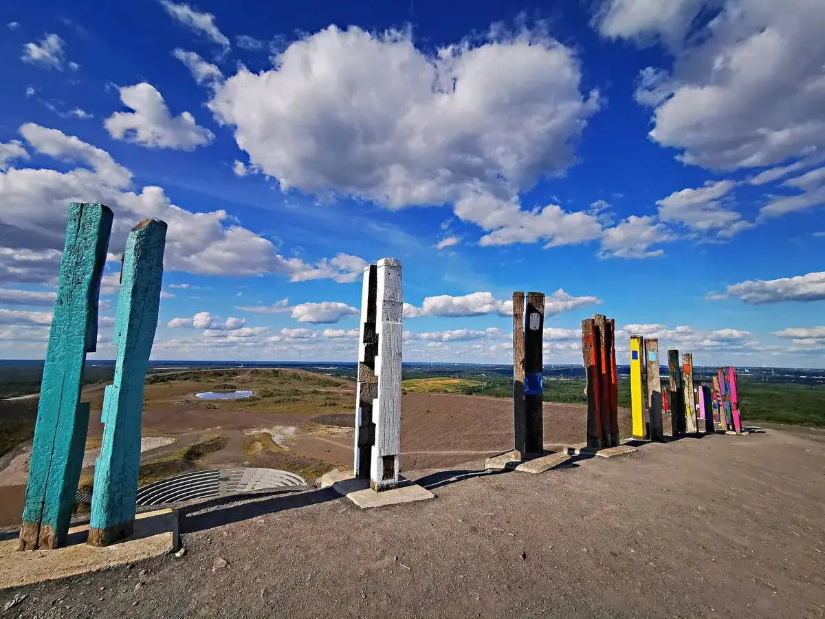 Bunte Holzskulpturen s&auml;umen den Weg auf der Halde Haniel. Der Himmel ist blau mit wei&szlig;en Wolken.