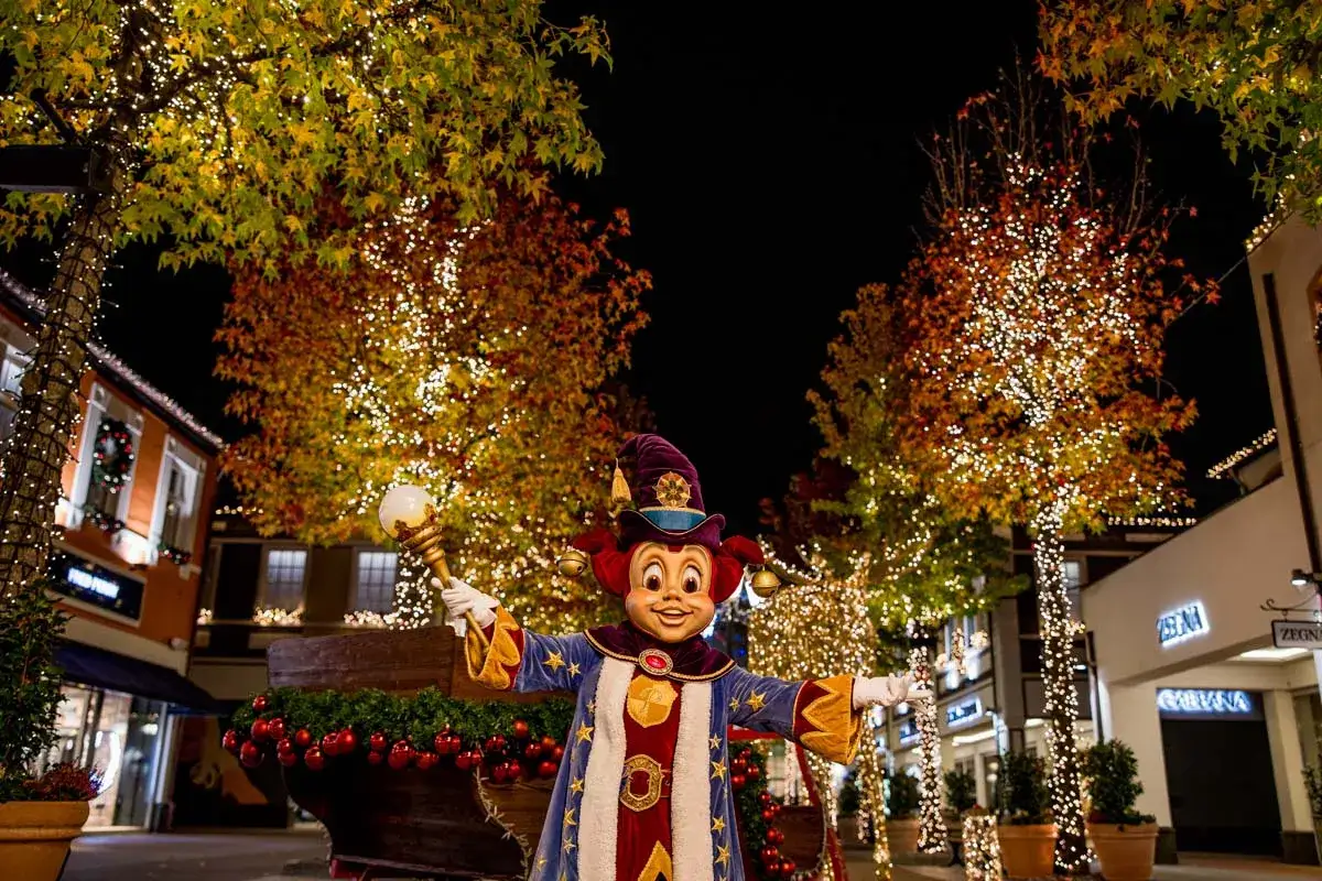 Ein fr&ouml;hlicher Clown mit Zauberstab steht vor B&auml;umen, die mit Lichterketten geschm&uuml;ckt sind. Der weihnachtsmarkt roermond ist magisch beleuchtet.