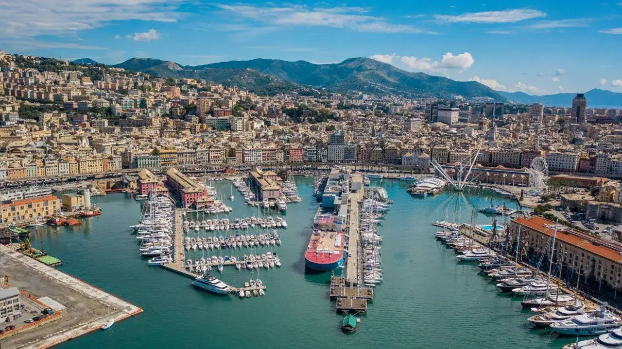 Blick auf den Hafen von Genua, Italien, mit vielen Yachten und einem großen roten Schiff. Die Stadt erstreckt sich bergauf hinter dem Hafen.