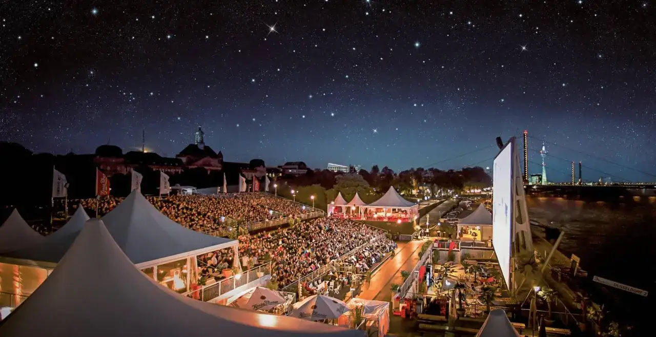 Ein belebtes Open-Air-Kino im Ruhrgebiet unter einem sternenklaren Himmel. Tausende Besucher sitzen auf Trib&uuml;nen, umgeben von Zelten und der Kulisse einer Stadt am Fluss.