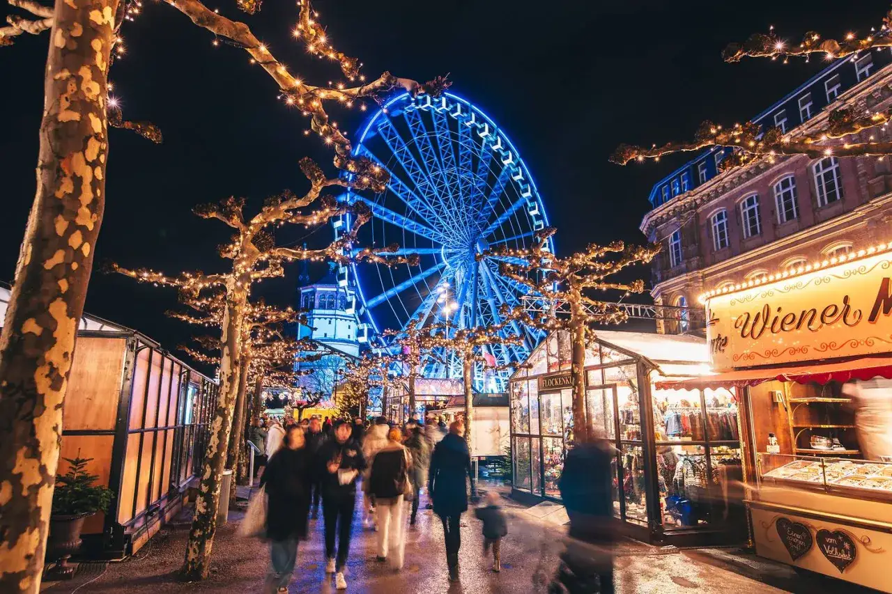 Menschen schlendern &uuml;ber einen stimmungsvollen Weihnachtsmarkt in NRW, beleuchtet von Lichterketten und einem Riesenrad. Ein echter Geheimtipp!