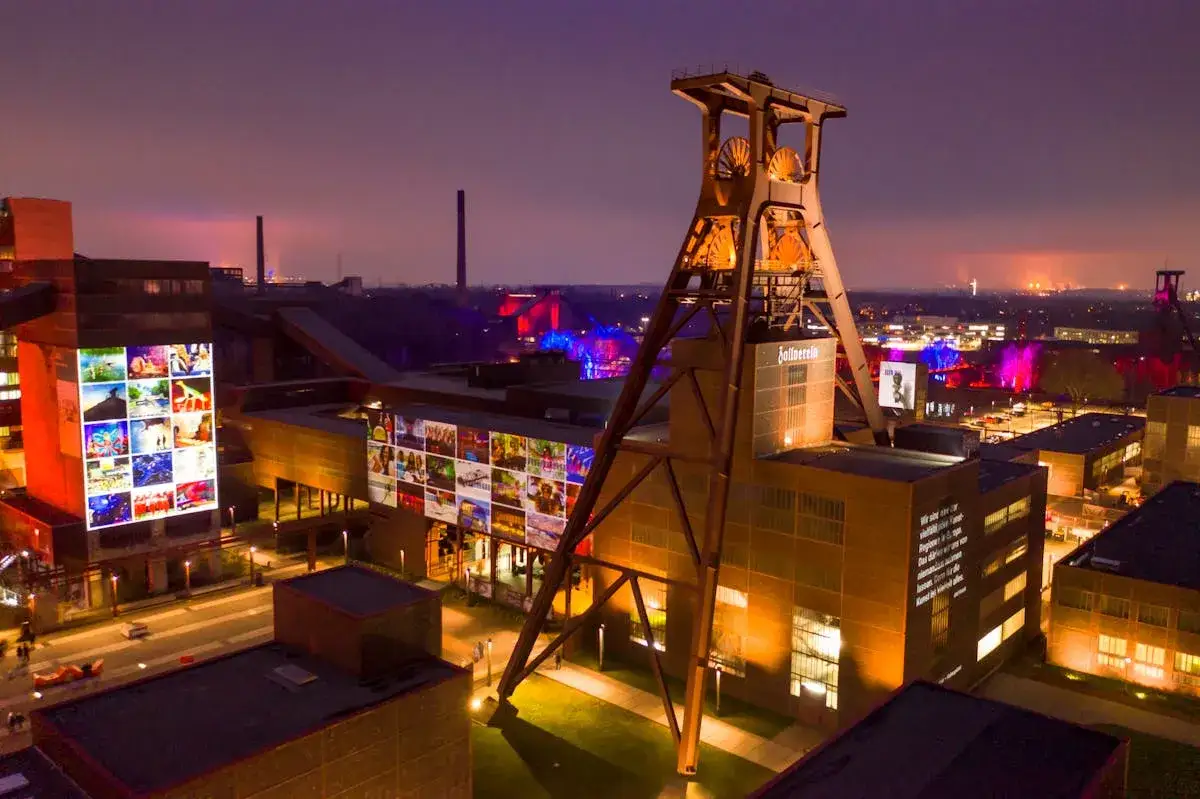 Die Zeche Zollverein nachts, beleuchtet mit bunten Projektionen auf den Geb&auml;uden und dem F&ouml;rderturm. Ein beeindruckendes Industriedenkmal im Abendlicht.