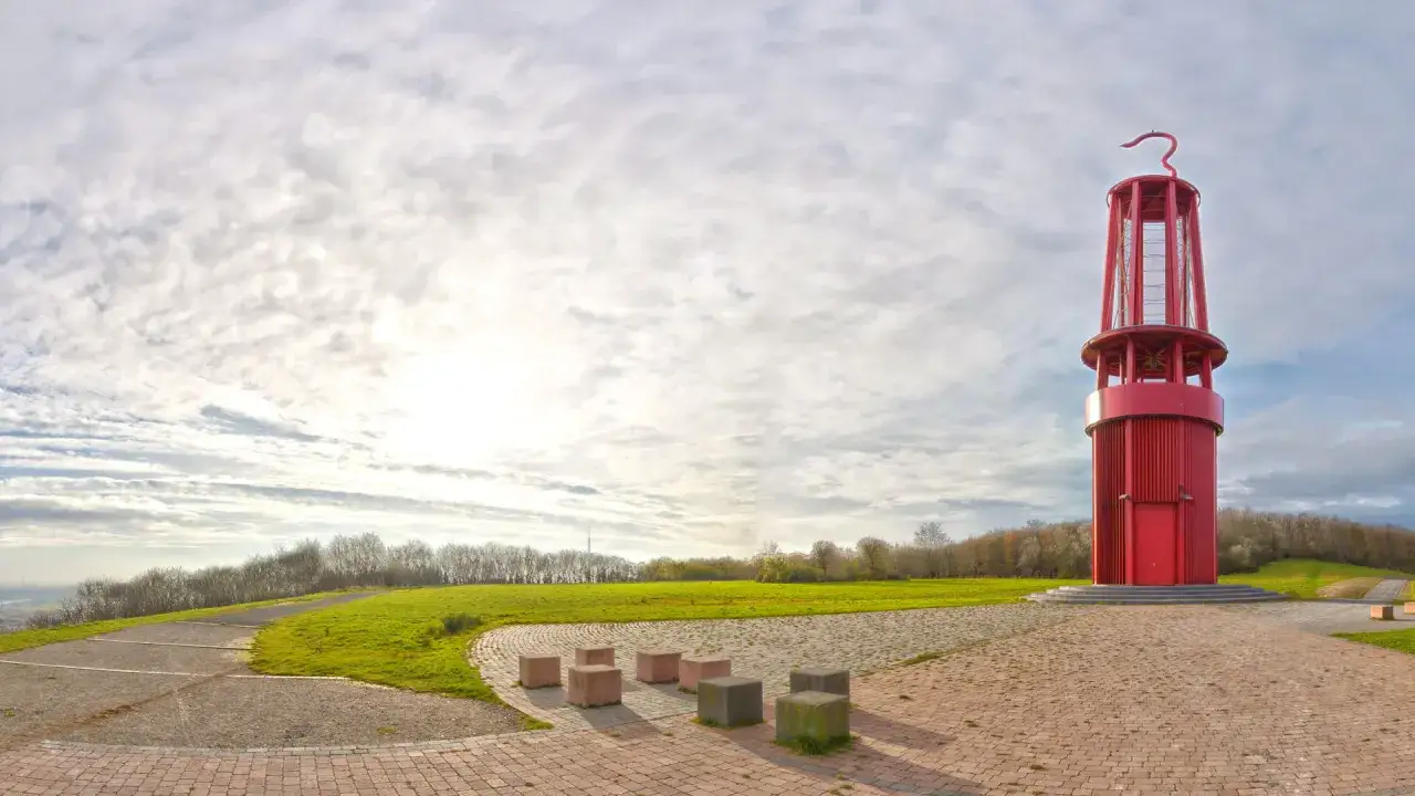 Rote Aussichtsturm auf der Halde Moers, umgeben von grünen Hügeln und einem weiten Himmel mit Wolken.