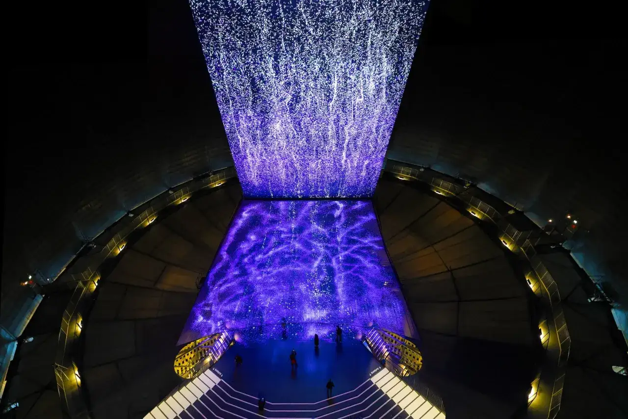 Imposante Lichtinstallation im Gasometer: Ein Wasserfall aus Sternen fällt auf eine violette, sternenklare Fläche. Besucher bestaunen das Spektakel.