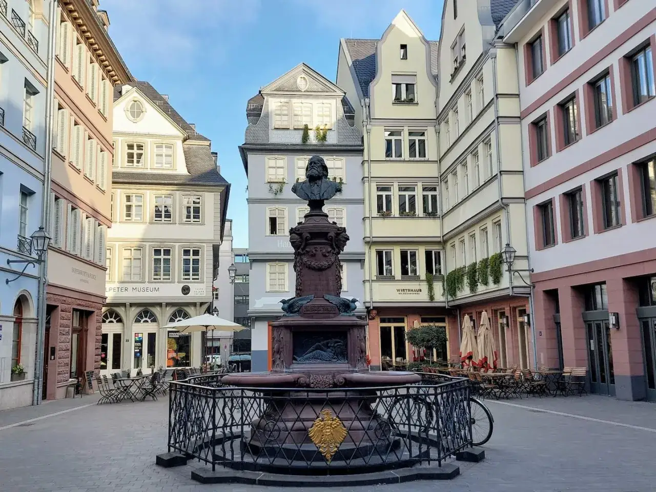 Brunnen mit B&uuml;ste im Zentrum eines Platzes in Frankfurt. Bunte H&auml;userfassaden und Stra&szlig;encaf&eacute;s s&auml;umen den Markt.
