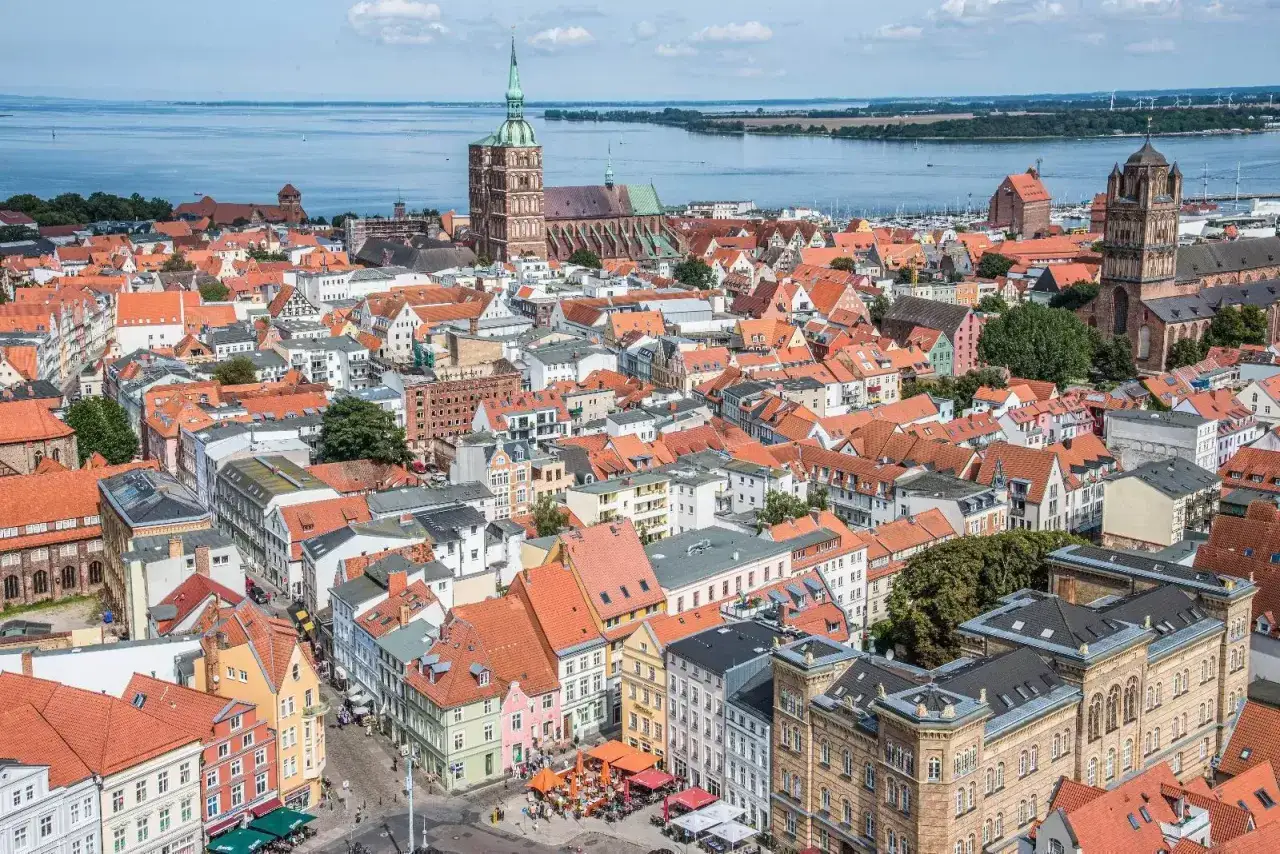 Blick über die roten Dächer der Stralsund Altstadt mit Kirchtürmen und dem Wasser im Hintergrund.