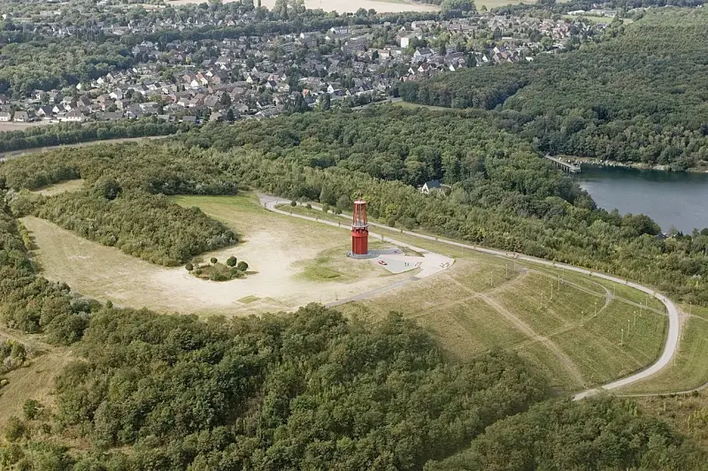 Luftaufnahme der Halde Rheinpreußen mit rotem Geleucht, umgeben von Wald und einem See.