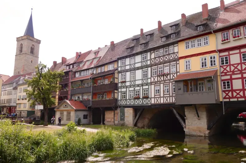 Fachwerkhäuser reihen sich am Wasser entlang, ein Kirchturm ragt empor. Ein idyllischer Anblick, um Erfurt entdecken zu gehen.